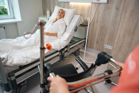 Foto de El paciente con una bata de hospital se sienta en una cama especial, junto a una enfermera con una silla de ruedas - Imagen libre de derechos