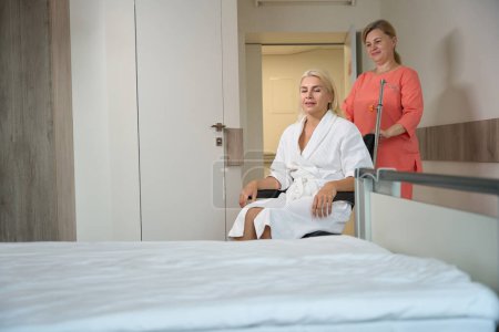 Foto de Enfermera cuidadora trajo a un paciente a la sala en una silla de ruedas, una mujer en una bata de hospital - Imagen libre de derechos