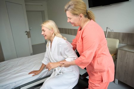 Téléchargez les photos : Genre travailleur de la santé aide dame transfert de fauteuil roulant au lit, femme en robe d'hôpital - en image libre de droit