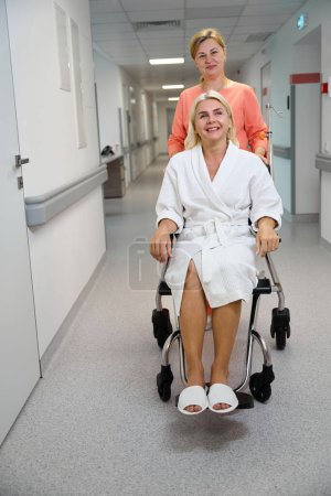 Foto de Empleado de la clínica lleva a una mujer sonriente en una silla de ruedas a lo largo del pasillo del hospital, la habitación está limpia y brillante - Imagen libre de derechos