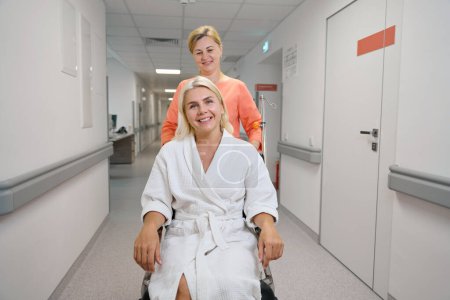 Foto de Trabajador de la salud lleva a una hermosa mujer en una silla de ruedas a lo largo de un pasillo del hospital, las mujeres se comunican dulcemente - Imagen libre de derechos