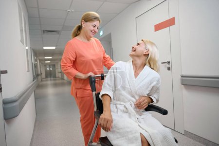 Foto de Trabajador de la salud lleva a un paciente en silla de ruedas a lo largo de un pasillo del hospital, las mujeres se comunican muy bien - Imagen libre de derechos