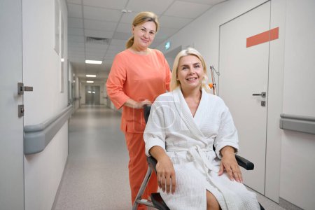 Foto de Amable enfermera lleva a un paciente en una silla de ruedas a lo largo del pasillo del hospital, la habitación está limpia y brillante - Imagen libre de derechos