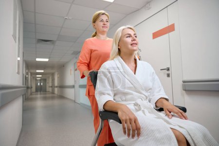 Téléchargez les photos : Une infirmière soignante emmène un patient en fauteuil roulant le long d'un couloir de l'hôpital, la chambre est propre et lumineuse - en image libre de droit