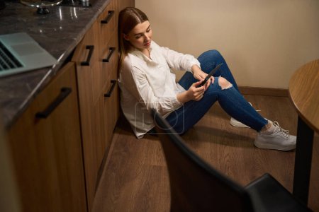 Téléchargez les photos : Femme réfléchie avec les cheveux longs dans des vêtements décontractés assis sur le sol et regardant couteau à l'intérieur - en image libre de droit
