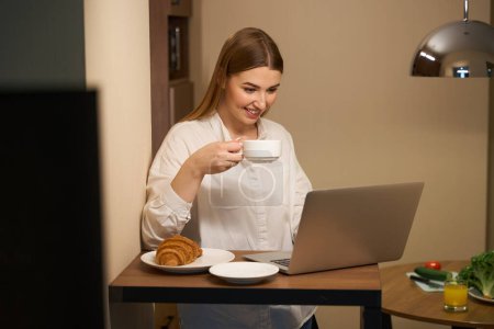 Photo for Smiling woman in casual clothes browsing laptop while snacking and standing in kitchen - Royalty Free Image
