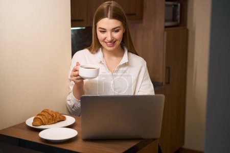 Photo for Smiling woman in casual clothes browsing notebook computer while drinking coffee and eating croissant indoors - Royalty Free Image