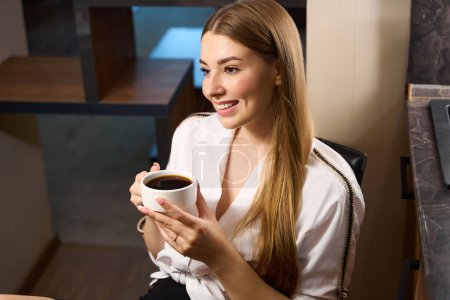 Foto de Mujer sonriente con hermosa manicura y cabello largo bebiendo café mientras está sentada en la silla en el interior - Imagen libre de derechos