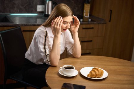 Foto de Mujer joven pensando mientras está sentado en la mesa en la que hay una taza de café y croissant en el interior - Imagen libre de derechos