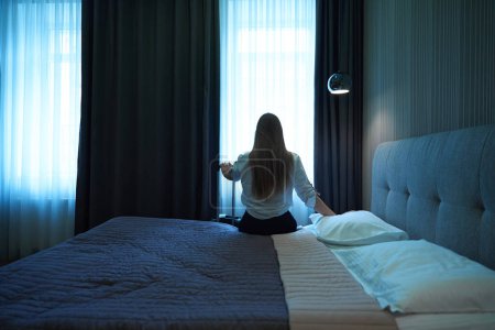 Foto de Vista trasera de la mujer caucásica con el pelo largo mirando por la ventana mientras está sentado en la cama en el hotel - Imagen libre de derechos