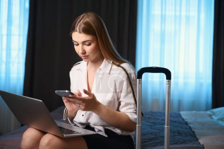 Téléchargez les photos : Belle femme avec ordinateur portable sur les genoux en utilisant mobile tout en étant assis sur le lit à l'hôtel - en image libre de droit