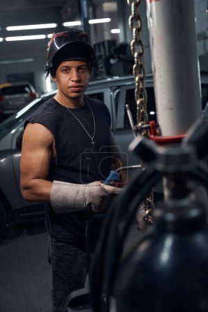 Foto de Hombre masculino guapo afroamericano trabajador de la estación de servicio de vehículos de motor que usa guantes protectores y mascarilla facial que trabaja con soldadura, reparación y soldadura de automóviles - Imagen libre de derechos