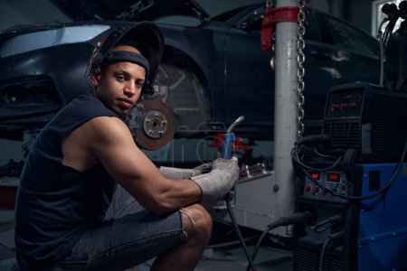 Photo for High-qualified man welder holding bar and adjusting welding machine before starting work with rusty and rotten car, wearing protective helmet and gloves, auto repair shop - Royalty Free Image