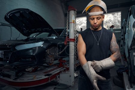 Photo for Focused African American auto-mechanic wearing welding gauntlets and protective helmet, going to weld rusty places on car, motor vehicle service station - Royalty Free Image