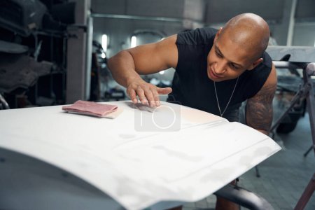 Photo for African American worker of car service station carefully removing wrinkles and ripples, sticking protective paper to part of car before painting - Royalty Free Image