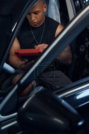 Téléchargez les photos : Auto-mécanicien afro-américain sérieux assis sur le siège du conducteur de la voiture et de programmation à bord de l'ordinateur qui peut enregistrer, calculer et afficher toutes sortes de choses - en image libre de droit