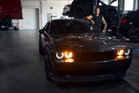 Téléchargez les photos : Homme afro-américain confiant debout près de voiture noire de luxe avec phares allumés, quittant la station-service de véhicules à moteur après réparation - en image libre de droit