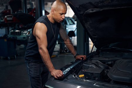 Photo for Masculine man worker of vehicle service station standing near luxury car with opened bonnet and looking inside, going to check oil level and add glass washer - Royalty Free Image