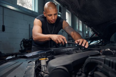 Photo for View to handsome bold auto-mechanic repairing and reconditioning car with opened bonnet using open-end spanner, motor vehicle service station - Royalty Free Image