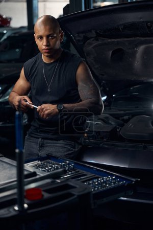 Photo for Attractive African American man with big tattoo on his hand changing sockets on socket wrench and looking at camera with self-confident expression, working at motor vehicle service station - Royalty Free Image