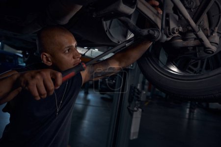 Photo for Focused African American auto-mechanic standing under the car and checking condition of car chassis, doing minor repairs using long screwdriver, automobile repair shop - Royalty Free Image