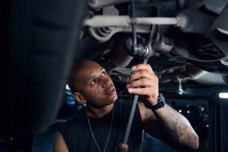 Photo for African American mechanic checking and repairing car axle, screwing spare parts with long screwdriver, adjusting or altering chassis standing under the car, auto service station - Royalty Free Image