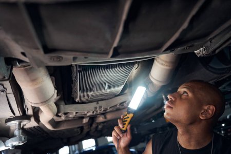 Photo for Attentive auto-mechanic carefully examining underbody of the car, checking condition of engine sump and exhaust pipes lighting with additional lamp, automobile repair shop - Royalty Free Image