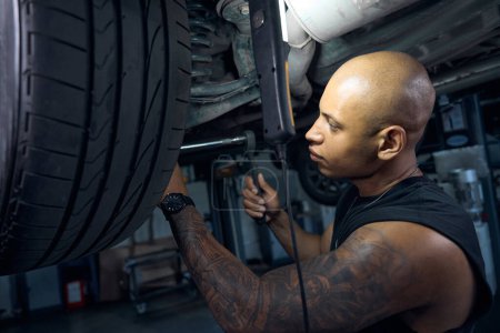 Photo for Focused African American man checking tightness and reliability of tightening the bolts and nuts on the car wheels using open-end spanner, auto service station - Royalty Free Image