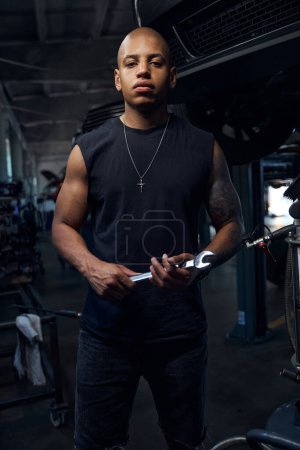 Photo for Self confident African American driver looking at camera holding wrench in hands, spending time in garage and repairing or tuning cars, auto repair shop - Royalty Free Image