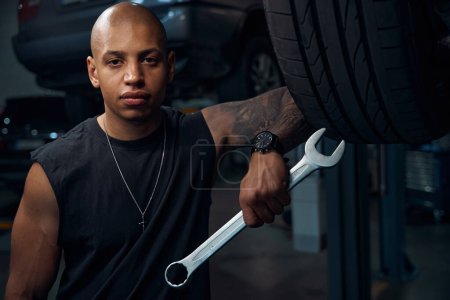 Photo for Handsome African American man auto-mechanic holding big socket wrench in hand, looking seriously, engaging in reconditioning and adjustment of any components of motor vehicle - Royalty Free Image