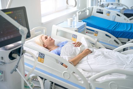 Téléchargez les photos : Femme d'âge moyen se trouve sur un lit d'hôpital spécial, elle est connectée à un goutte à goutte et une bouteille d'oxygène - en image libre de droit