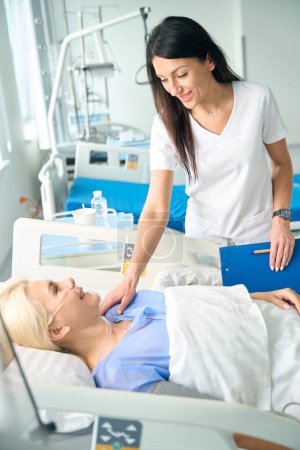 Foto de El médico anima a una mujer después de la cirugía, la paciente se acuesta en una cama postoperatoria especial - Imagen libre de derechos