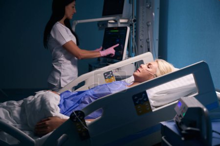 Foto de Recuperando rubia se encuentra en la sala de recuperación, una enfermera supervisa su condición utilizando equipos modernos - Imagen libre de derechos