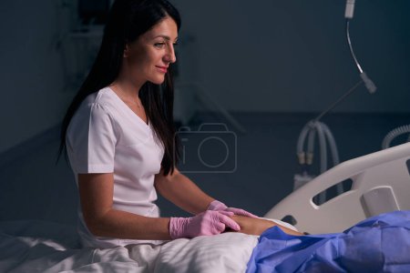 Foto de Trabajadora médica en guantes de protección se comunica con un paciente después de la cirugía, ella sostiene su mano - Imagen libre de derechos