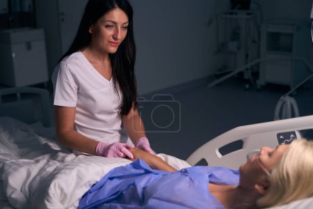 Foto de Joven médico en guantes de protección se comunica con un paciente después de la cirugía en una habitación del hospital, ella sostiene su mano - Imagen libre de derechos
