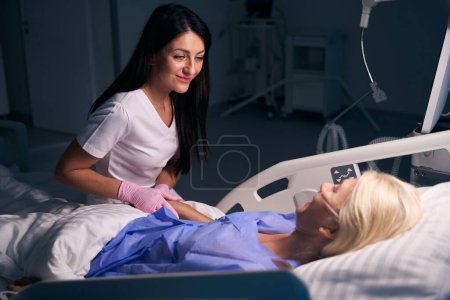 Foto de El joven médico se comunica con un paciente después de la cirugía en una habitación de hospital, ella sostiene su mano - Imagen libre de derechos