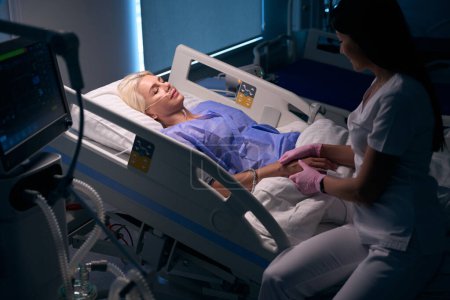 Foto de Enfermera se comunica con un paciente después de la cirugía en una habitación de hospital, ella sostiene su mano - Imagen libre de derechos