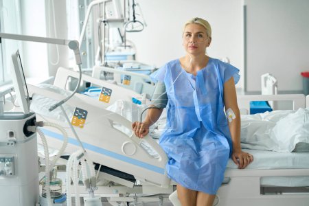 Señora en bata de hospital se sienta en la cama en la unidad de cuidados intensivos, ella está conectada a máquinas de apoyo y monitoreo