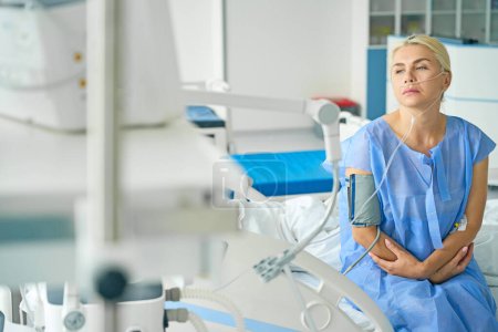 Foto de Mujer triste se sienta en una cama de hospital en cuidados intensivos, ella está conectada a un tonómetro y una máquina de oxígeno - Imagen libre de derechos