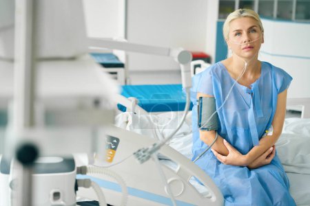 Téléchargez les photos : Femme fatiguée assise sur un lit d'hôpital spécial, elle est connectée à un tonomètre et à une machine à oxygène - en image libre de droit