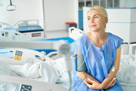 Téléchargez les photos : Femme fatiguée est assise sur un lit d'hôpital spécial, elle est connectée à un tensiomètre et à une machine à oxygène - en image libre de droit