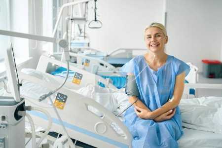 Foto de Rubia sonriente se sienta en una cama de hospital especial, ella está conectada a un monitor de presión arterial y una máquina de oxígeno - Imagen libre de derechos