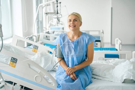 Téléchargez les photos : Blonde d'âge moyen assise sur un lit d'hôpital spécial, elle est connectée à un tensiomètre et à une machine à oxygène - en image libre de droit