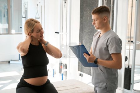 Téléchargez les photos : Femme enceinte se plaint à un physiothérapeute de douleur au cou, il écrit tout dans un dossier bleu - en image libre de droit