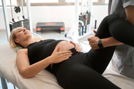 Téléchargez les photos : Mignonne femme enceinte à une séance de chiropratique confortable, spécialiste travaillant sur ses pieds - en image libre de droit