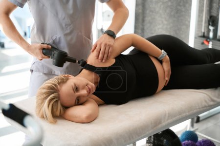 Téléchargez les photos : Le physiothérapeute utilise un appareil de physiothérapie spécial, tandis qu'une cliente enceinte s'allonge sur une table de massage - en image libre de droit