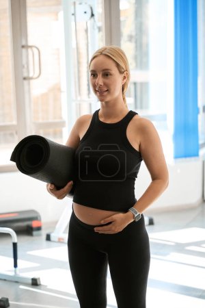 Téléchargez les photos : Femme blonde enceinte dans la salle de gym tient un tapis de gymnastique roulé dans un rouleau, il ya beaucoup de lumière du soleil à l'intérieur - en image libre de droit