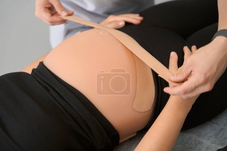 Téléchargez les photos : Spécialiste kinésithérapeute tape le ventre d'une femme enceinte, le client s'allonge sur la table de massage - en image libre de droit