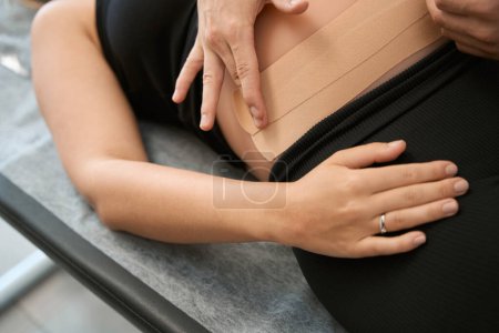 Foto de Especialista en rehabilitación toca el vientre de una mujer embarazada, el cliente se acuesta en la mesa de masaje - Imagen libre de derechos