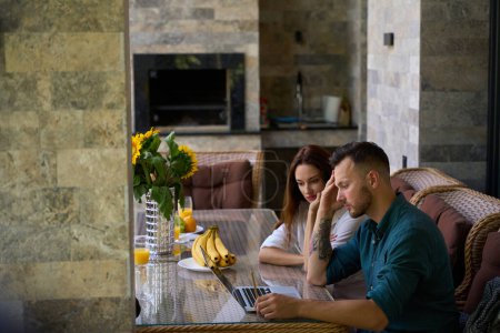 Photo for Young man works on the terrace at his laptop, next to his beautiful wife - Royalty Free Image